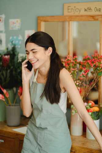 A florist in an apron laughs while taking a phone call at the counter of her flower shop, surrounded by fresh flowers.