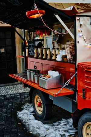 A red food truck with string lights parked on a cobblestone street, serving food through its window in a wintertime outdoor setting